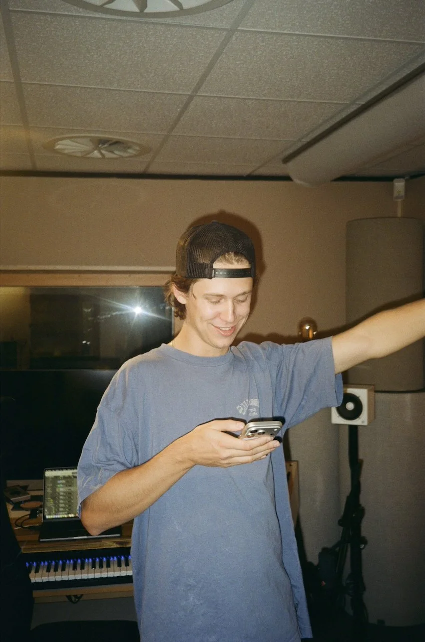 Junge Person in einem Tonstudio benutzt ein Smartphone, trägt einen Baseballcap rückwärts, im Hintergrund sind Musikgeräte und Lautsprecher sichtbar.