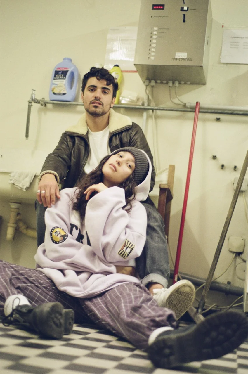 Ein Mann sitzt hinter einer sitzenden jungen Frau auf dem Boden in einem Raum, der wie eine Waschküche aussieht, mit Waschmittel im Hintergrund.