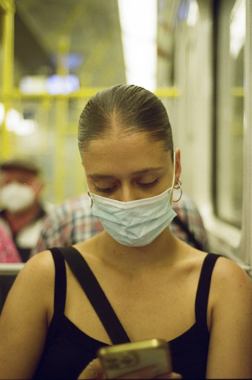 Eine junge Frau mit einem schwarzen Top trägt eine medizinische Maske und schaut auf ihr Smartphone, während sie in einem öffentlichen Nahverkehrsmittel sitzt. Im Hintergrund sind weitere Passagiere ebenfalls mit Masken zu sehen.
