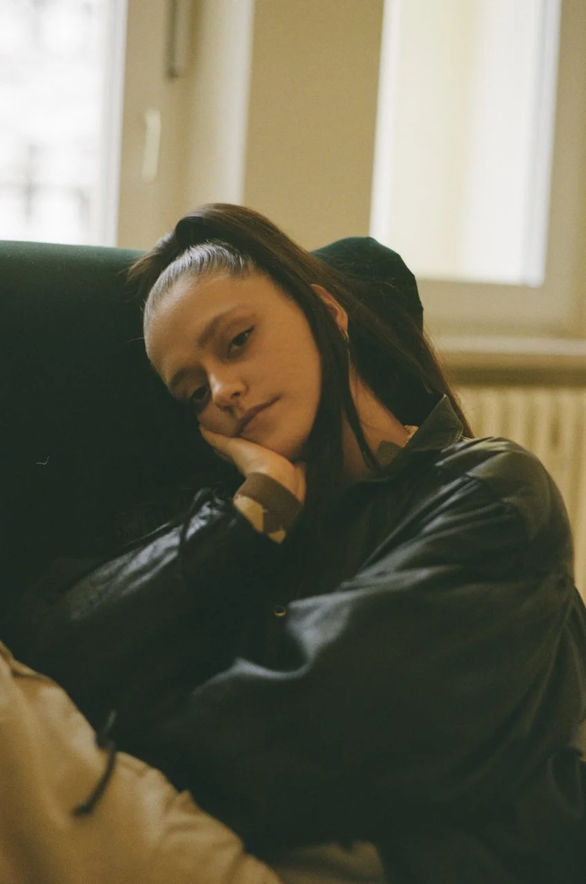 Eine junge Frau sitzt nachdenklich an einem Fenster, ruht ihren Kopf auf der Hand und trägt eine schwarze Jacke.