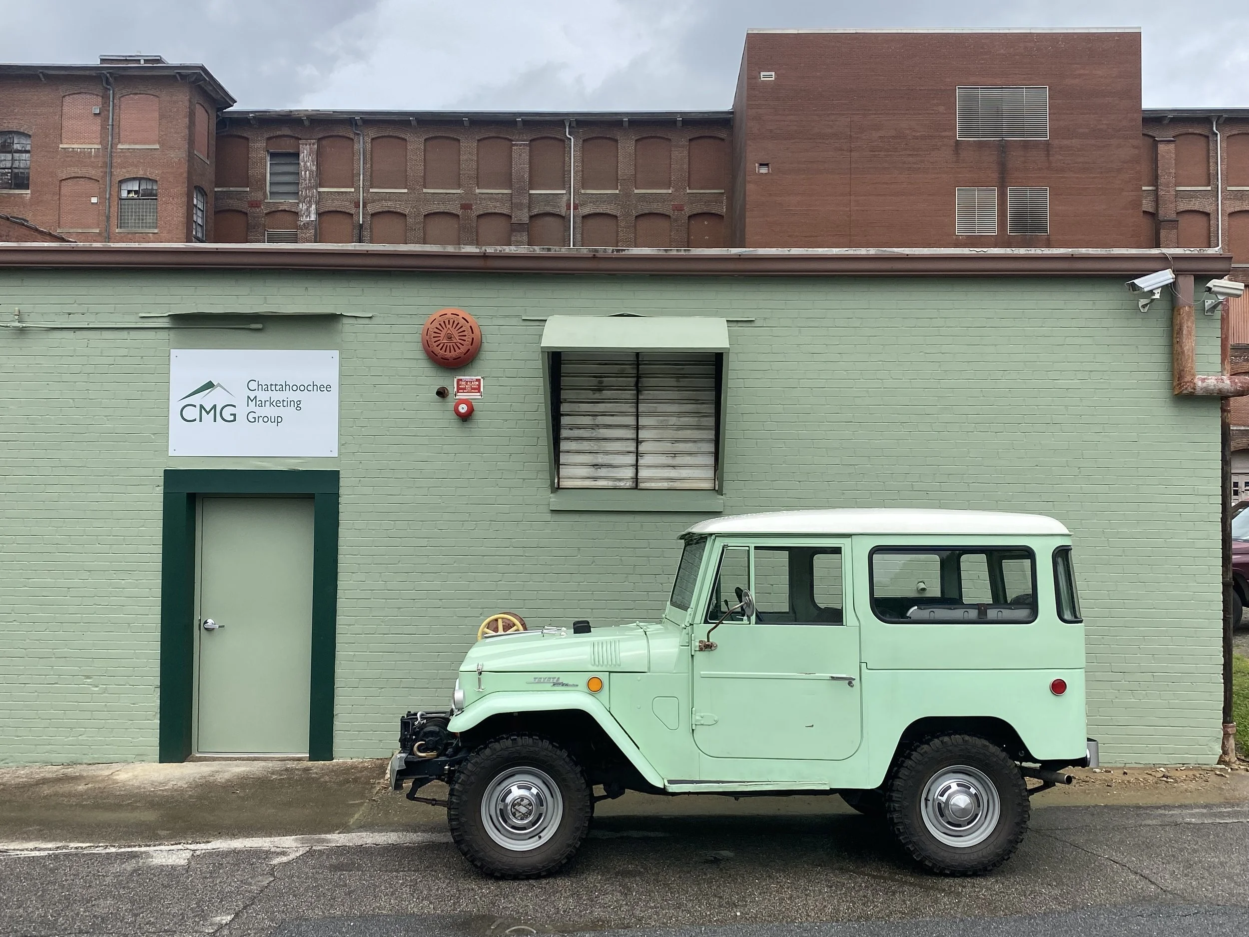A vintage mint green Toyota Land Cruiser parked in front of a pale green brick building with a sign that reads 'Chattahoochee Marketing Group.' The building has a window, a red fire alarm, and a green door, with a large brown brick building in the background.