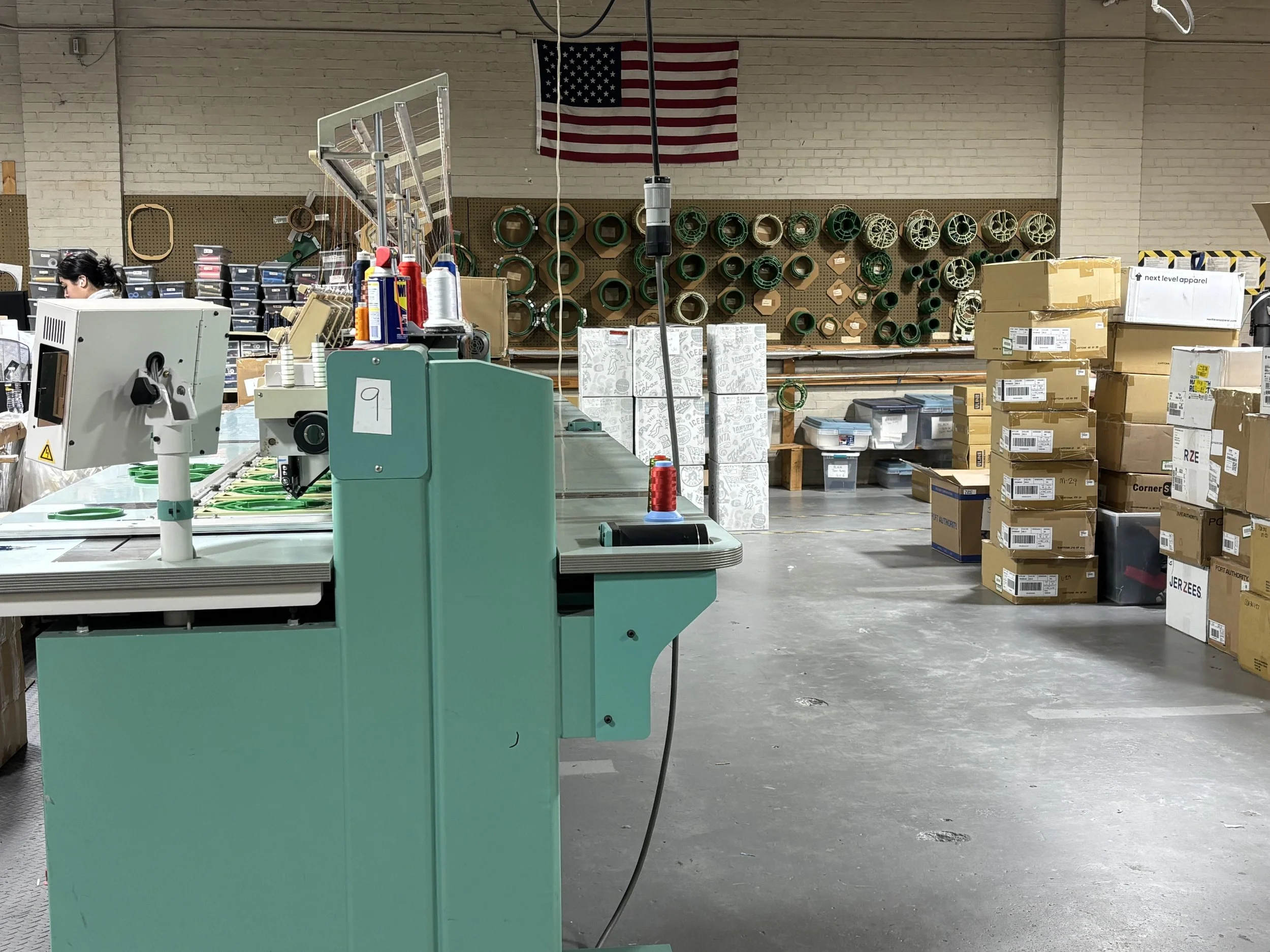 A workspace inside a warehouse, with stacked boxes, cardboard cartons, and organized shelves of spools and rolls. An American flag hangs on the wall, with a cutting or printing machine in the foreground.