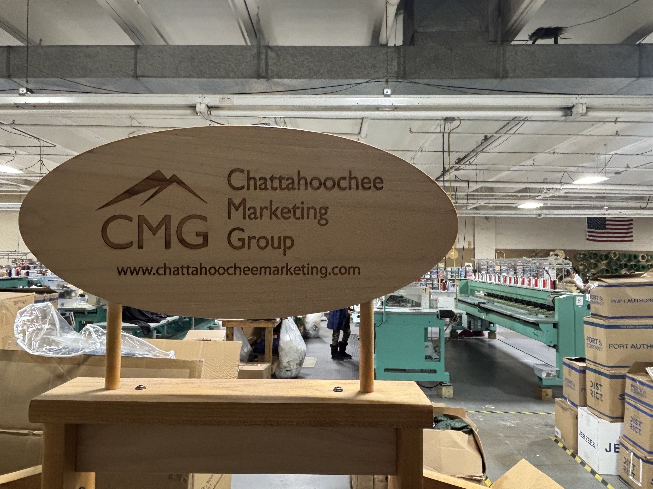 Wooden sign for Chattahoochee Marketing Group displaying logo and website, with an industrial warehouse background containing boxes, conveyor belt, and workers.