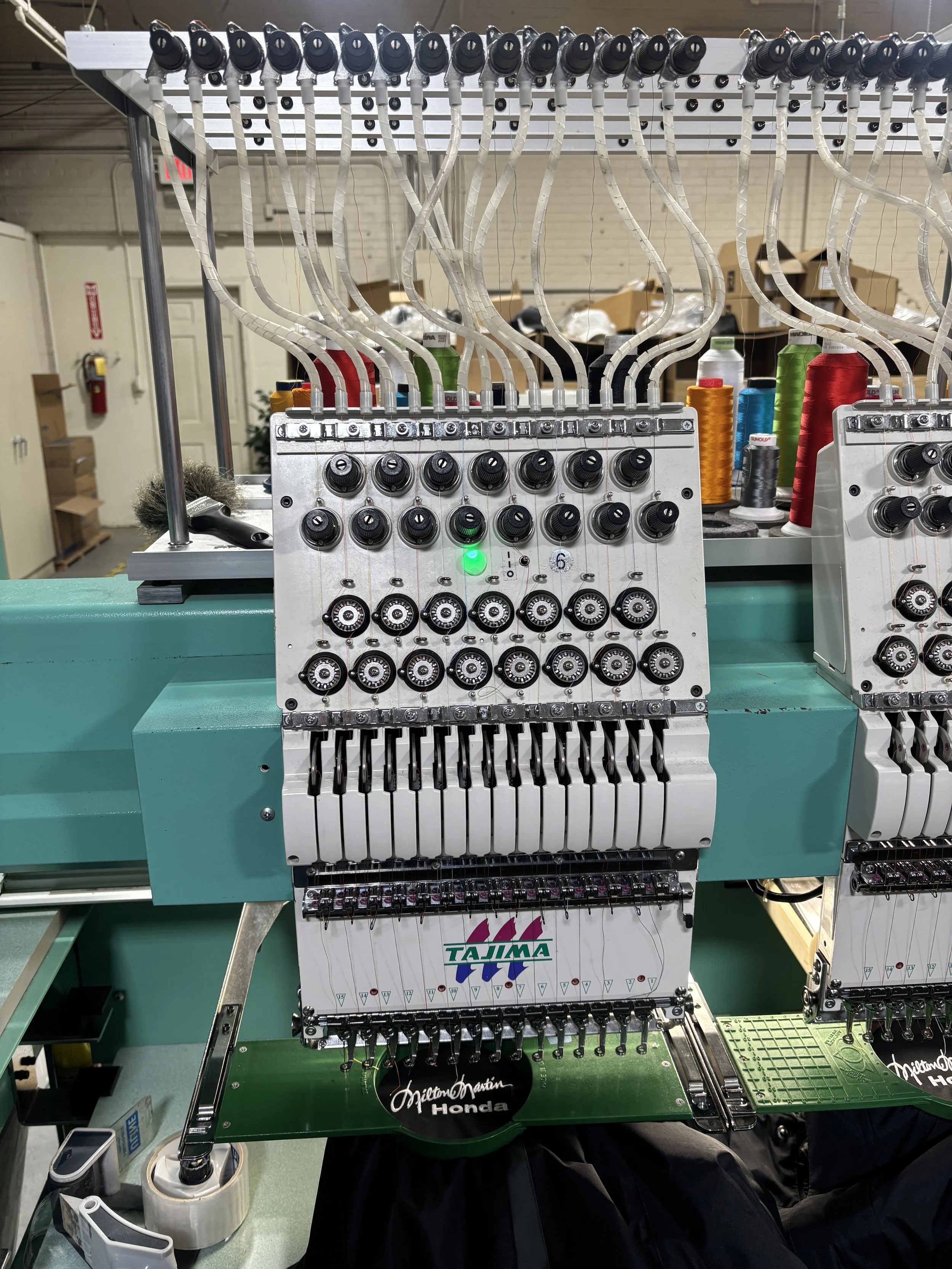 A close-up view of a multi-thread embroidery machine with several colored spools of thread in the background, used for sewing designs onto fabrics.