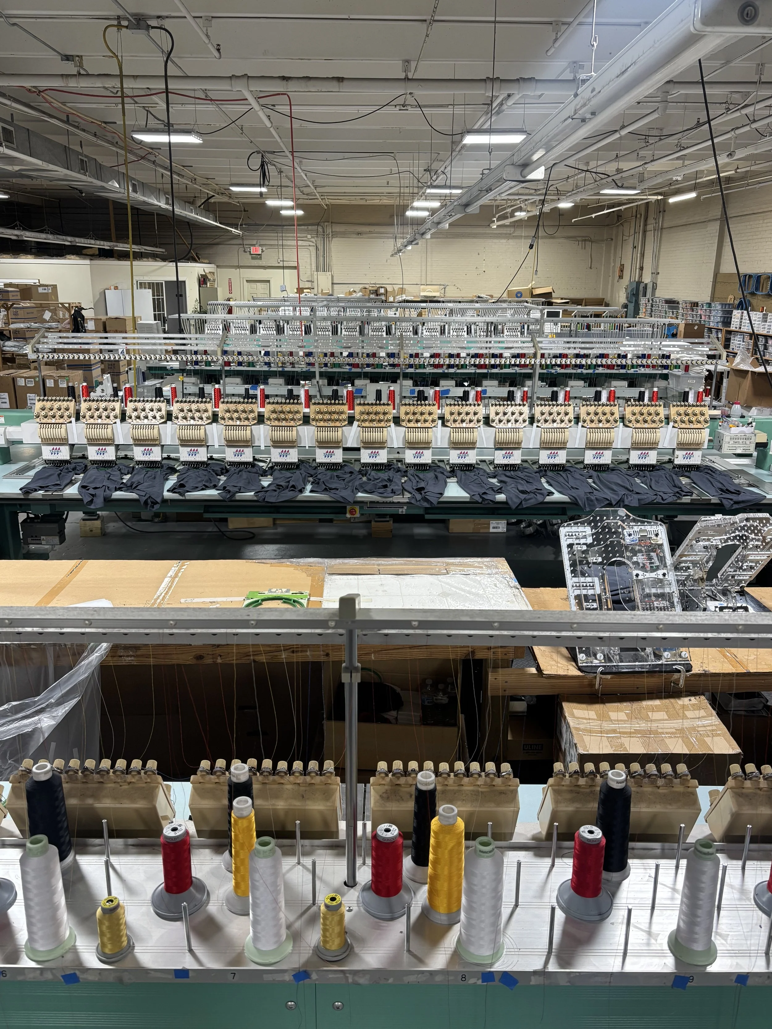 Industrial textile sewing machines with black fabric being stitched, spools of thread in various colors in the foreground, and an organized workshop with boxes and equipment in the background.