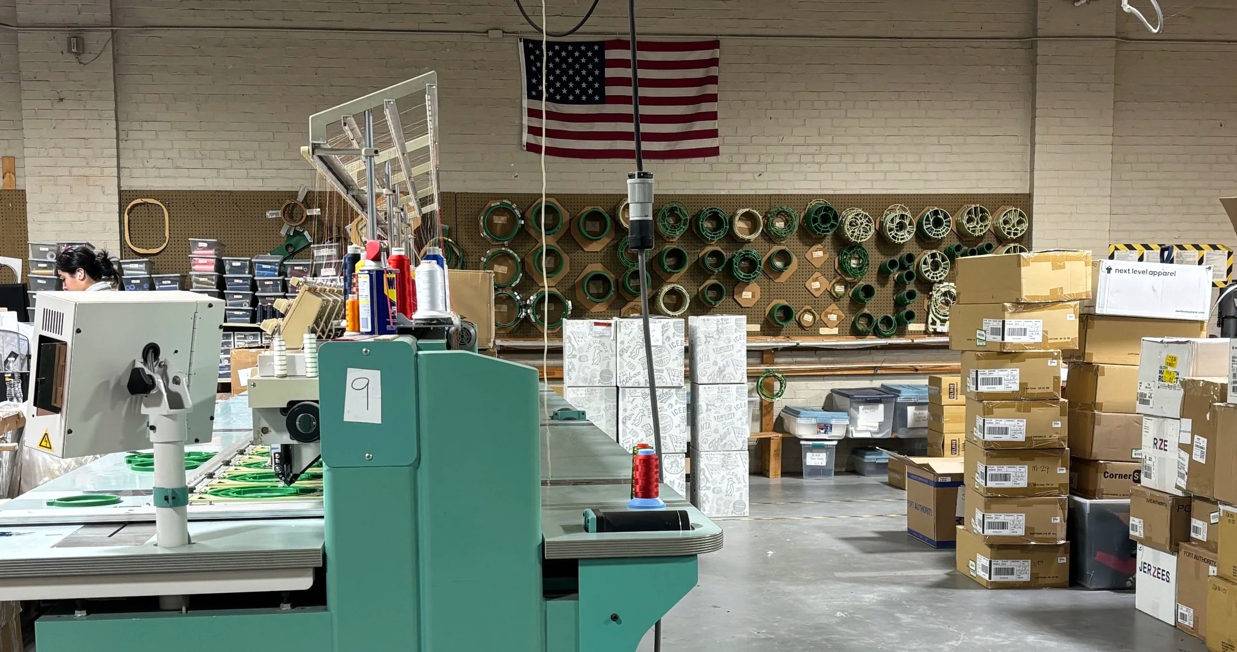 Warehouse or factory workspace with cardboard boxes, plastic containers, and shelves filled with products. There is a sewing machine, spools of thread, and packaging materials visible. An American flag hangs on the wall in the background.