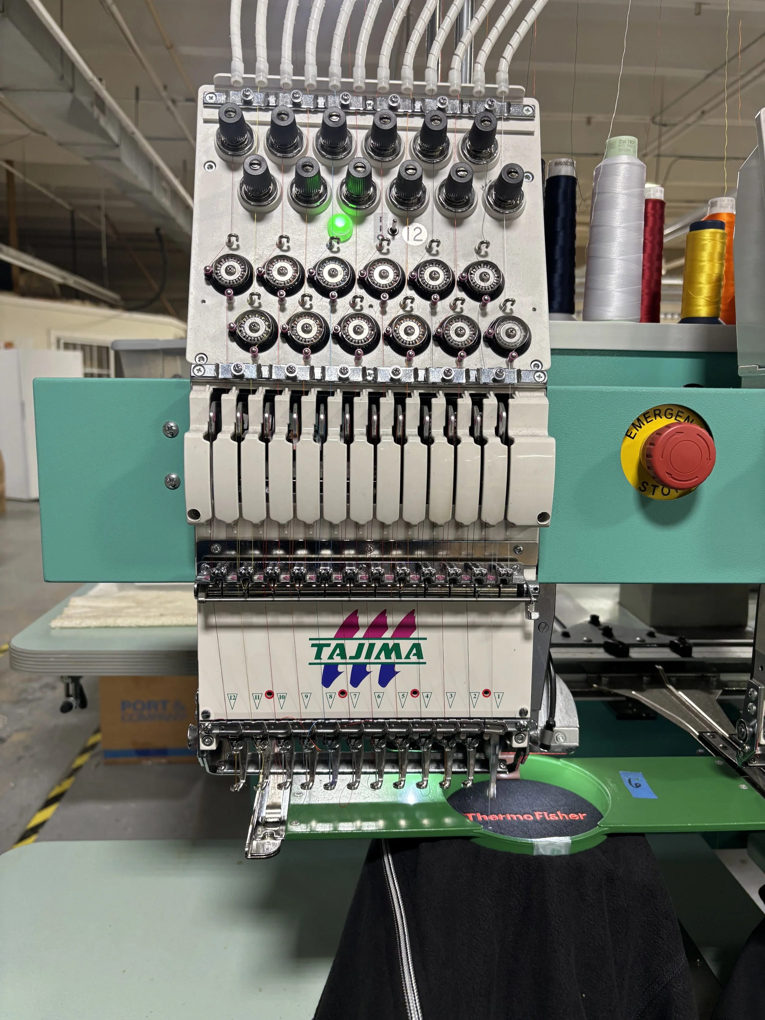 Close-up of an industrial embroidery machine with spools of thread in various colors to the right, and the brand name Tajima on the lower part. The machine has multiple needle heads and a control system with a green indicator light.