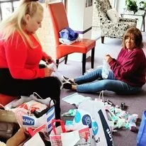 Two women sitting on the floor surrounded by various household and personal items, including bags and clothing, in a living room.