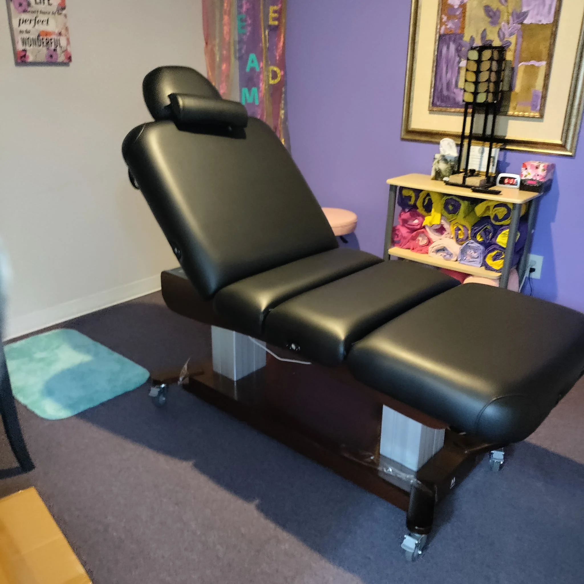 Black massage or chiropractic adjustment table with adjustable headrest in a room with purple walls, decorated with colorful artwork, a small table with rolled towels, a lamp, and various decorative items.