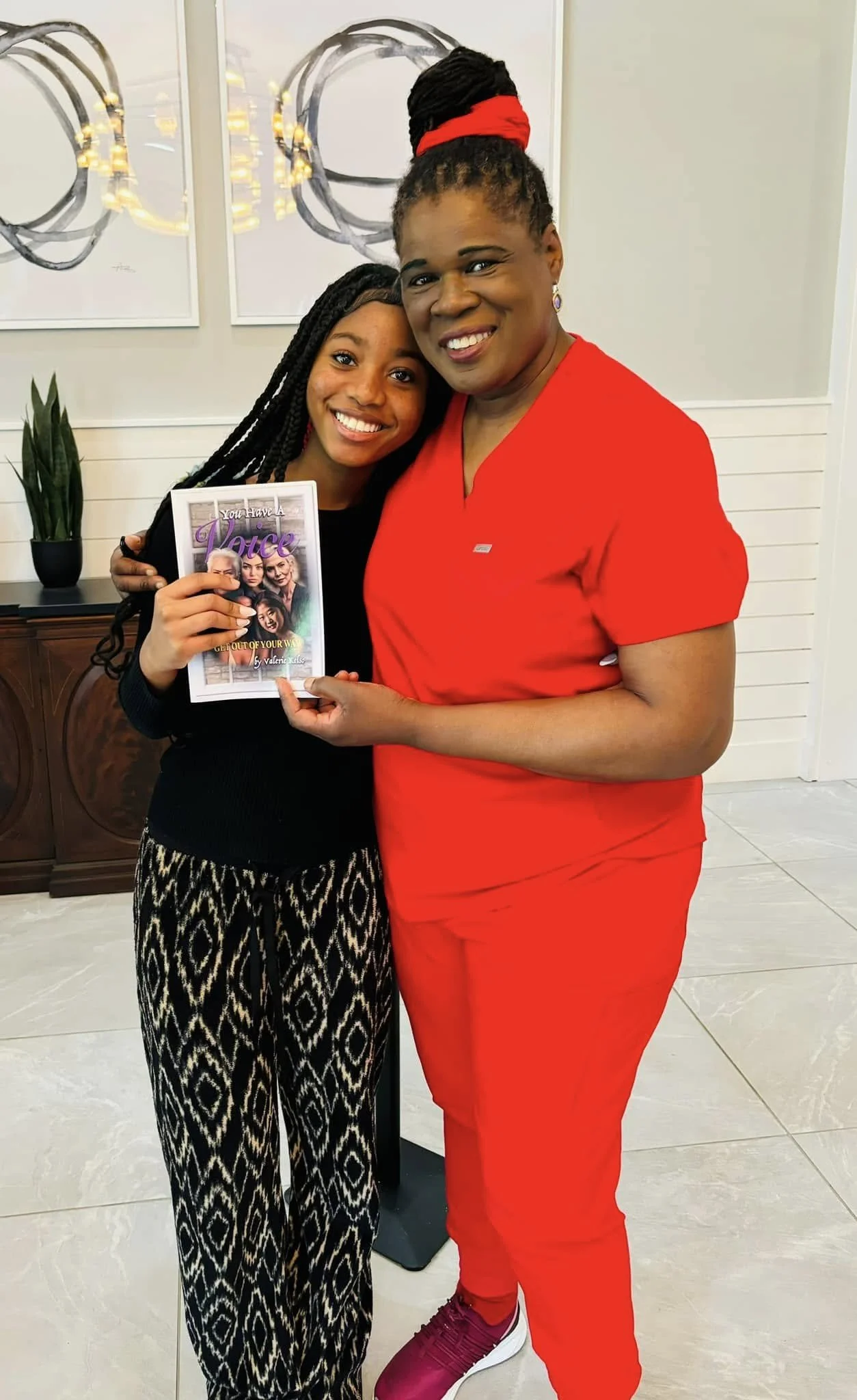 Two women smiling and posing together, one holding a book titled "You Have A Voice, Get Out Of Your Way". The woman on the right wears a bright red outfit and the woman on the left has patterned pants and a black top.