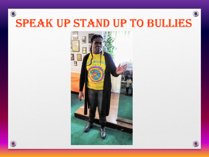 A woman standing indoors, wearing a yellow T-shirt with a 'Stop Bullying' message, black pants, and a black coat. She is making a gesture with her hand, and the background includes a mirror, a plant, and some pictures on the wall. The image promotes 