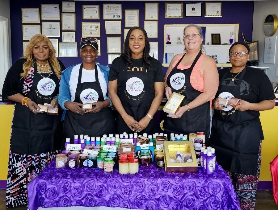 Five women standing behind a table filled with various bottles and boxes of products, in a room with framed certificates on the wall, all wearing aprons with a logo. They are smiling and holding some of the products.