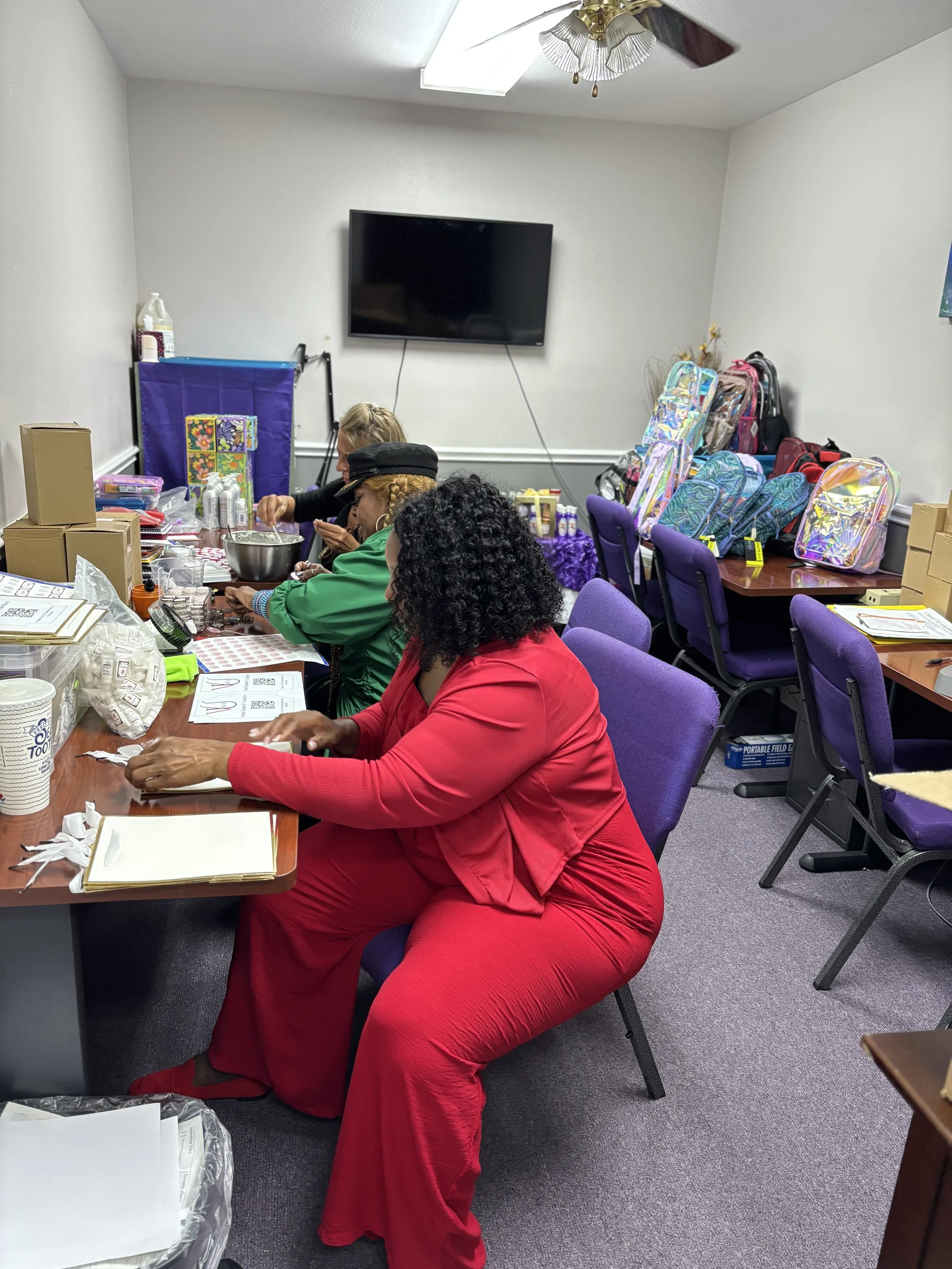 Women sitting at a table filling out paperwork in a room with backpacks and gift bags on the tables and chairs.