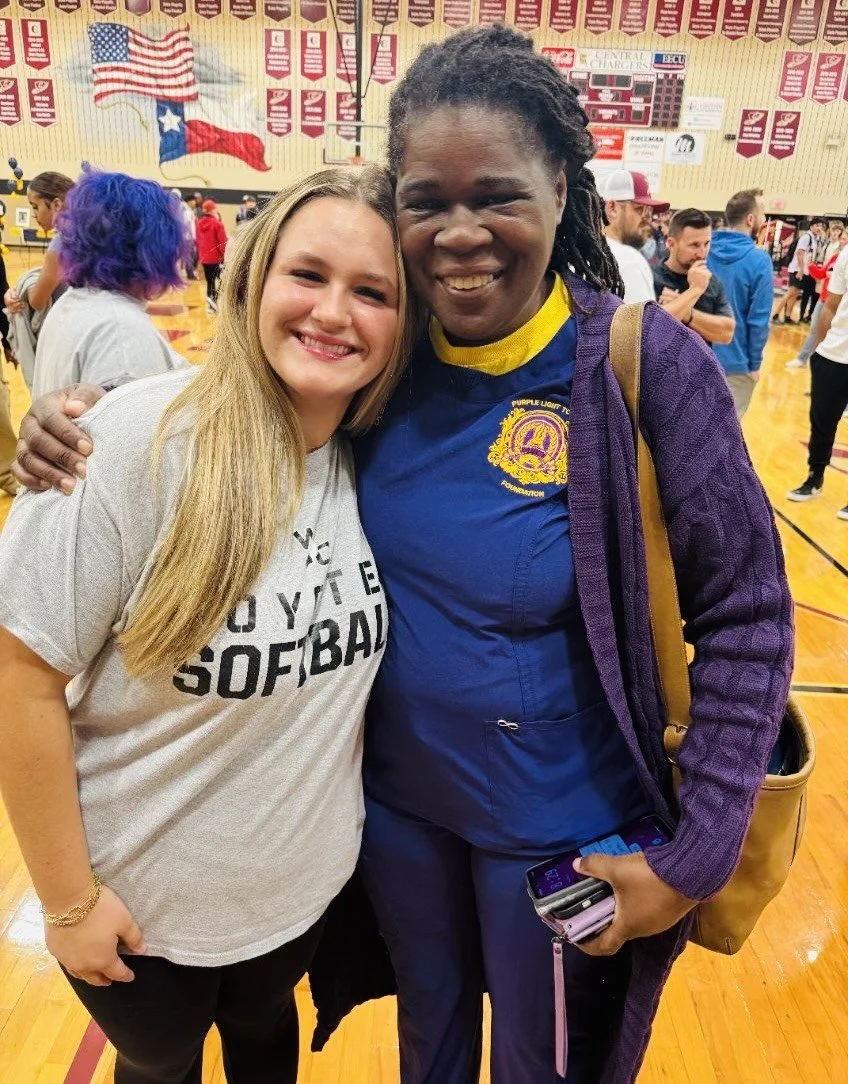 Two women smiling and hugging inside a gymnasium with banners and flags hanging from the ceiling. One woman has long blonde hair, and the other has short dreadlocks and is wearing a purple jacket and a blue shirt with a yellow collar and emblem.