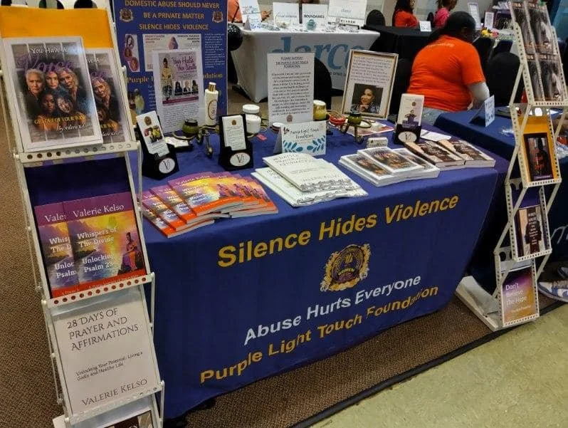 A booth with a table covered in a blue cloth that reads 'Silence Hides Violence,' and 'Abuse Hurts Everyone, Purple Light Touch Foundation.' The table displays books, pamphlets, candles, and framed information about domestic violence. There are liter