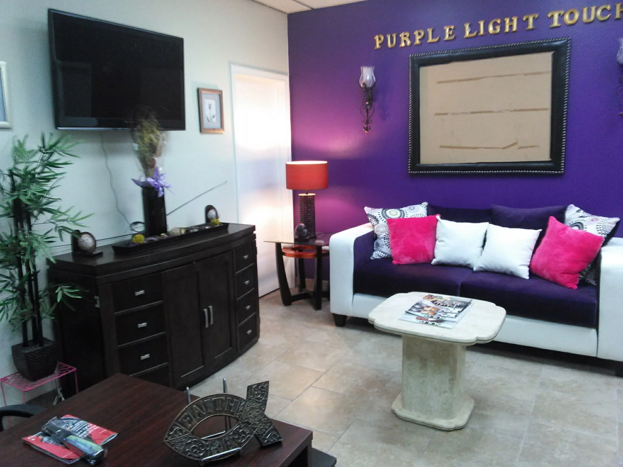 Living room with a purple accent wall, black and white sofa with pink and white cushions, a small beige table, a dark wood TV stand with decorations, a flat-screen TV, a tall potted plant, framed pictures, a side table with a lamp, and wall decor.