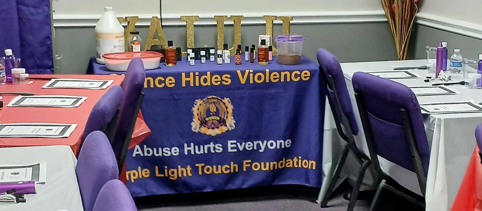 Table with printed informational sheets and purple chairs surrounding it, against a backdrop of a purple and orange table with bottles of massage oils, a container, and decorations, including a banner that reads 'Violence Hides Violence' and 'Abuse H