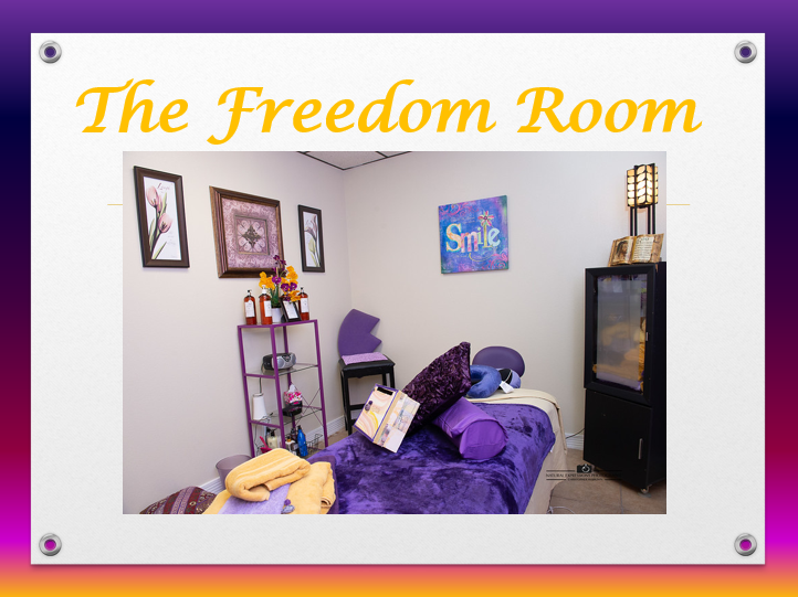 A cozy therapy or counseling room with a purple-themed bed and pillows, framed artwork on the walls, a small purple bookshelf with decorative items, a black cabinet, and a lit decorative lamp, labeled "The Freedom Room" at the top.