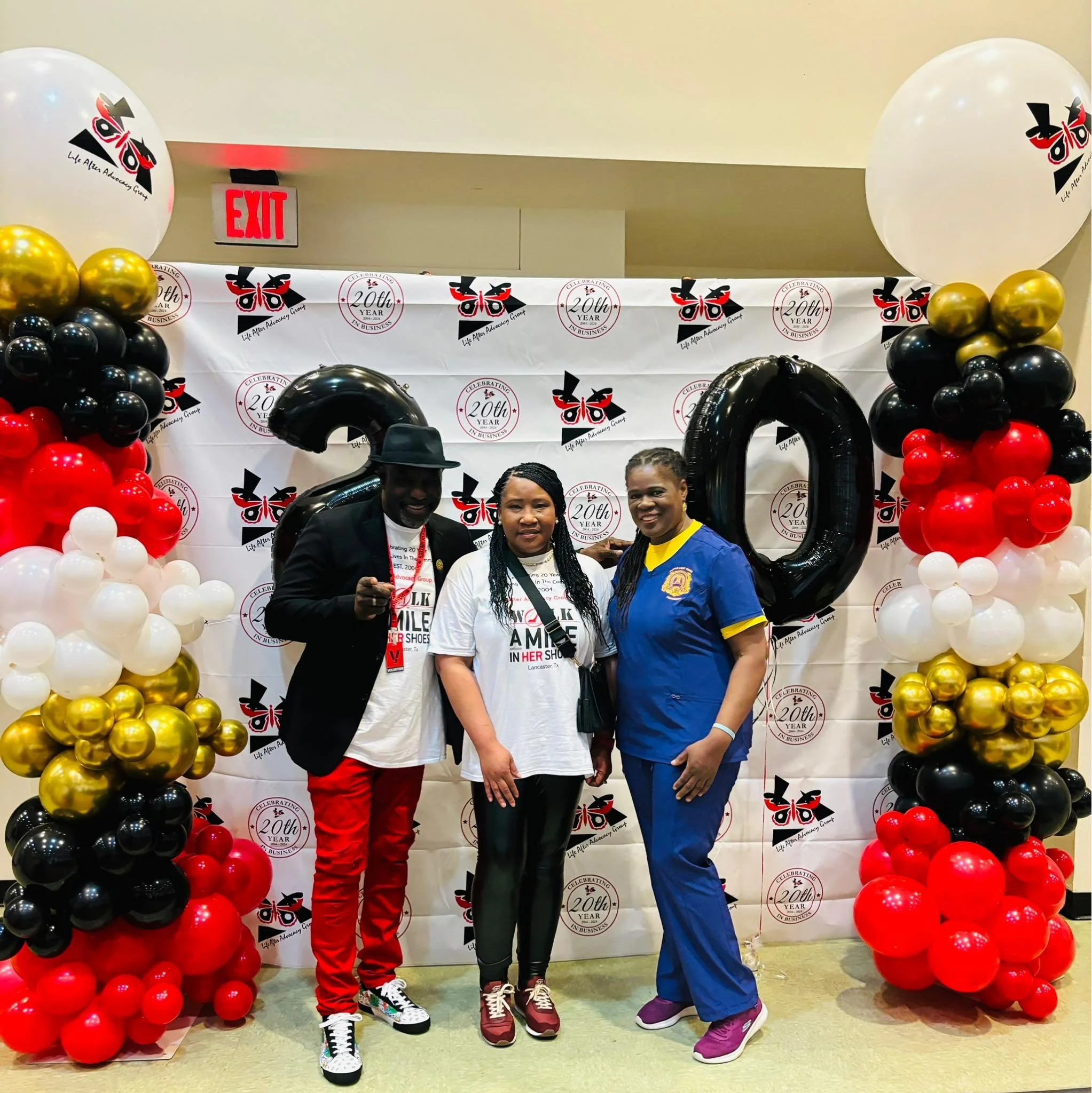 Three people standing in front of a celebration backdrop with balloons and the number 20. The person on the left is wearing a black hat, black blazer, white T-shirt, red pants, and sneakers. The person in the middle is wearing a white T-shirt and bla