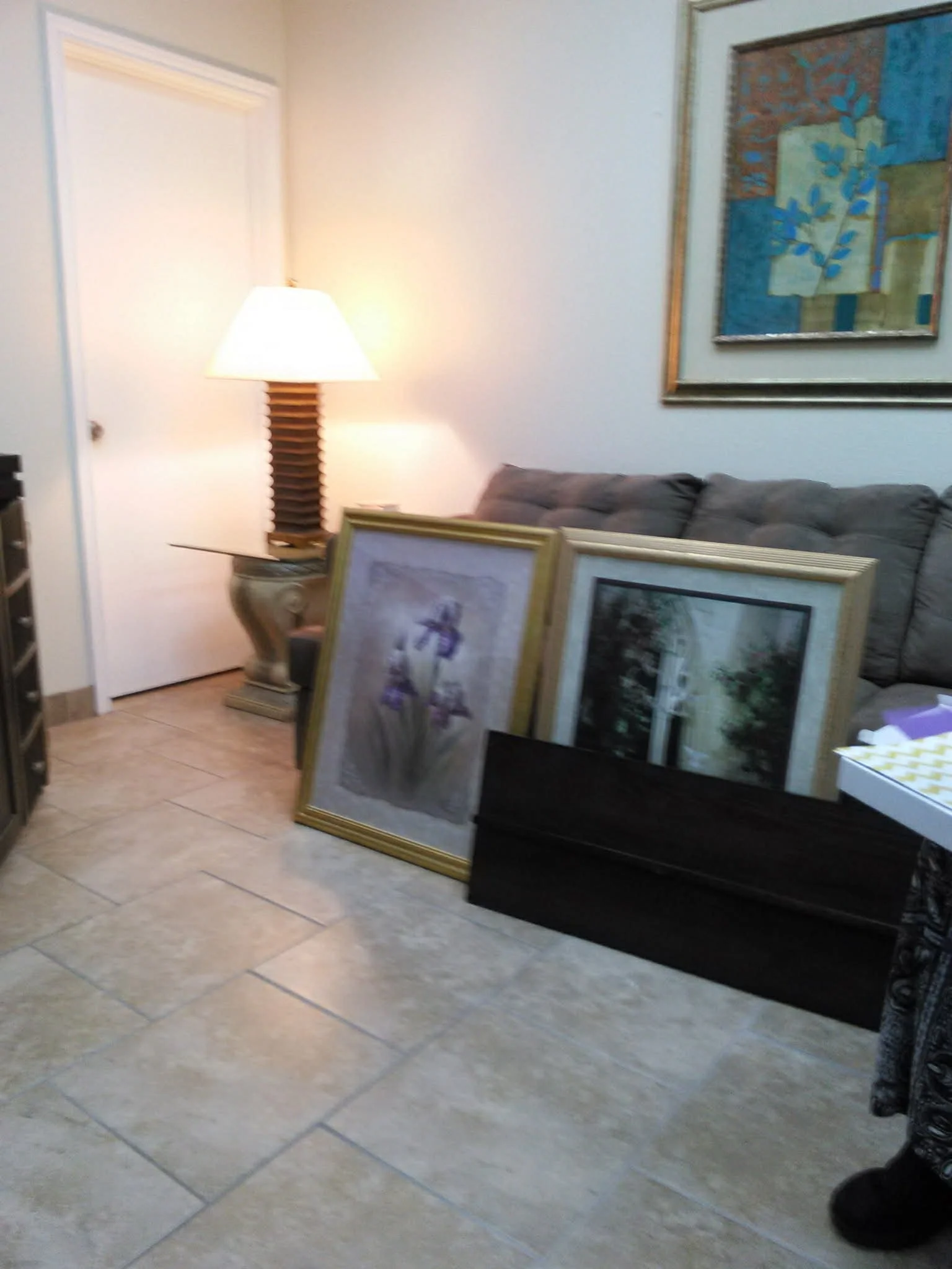 A living room with a sofa, a table lamp, framed pictures leaning against the sofa, an abstract painting on the wall, a door, and a tiled floor.