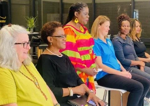 A diverse group of six women sitting in a line in a professional setting, with some smiling and others attentively listening.