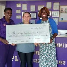 Three women standing together in an indoor setting, holding a large check, smiling at the camera.