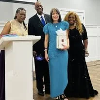 Four people standing together indoors, with one woman holding an award or certificate, smiling.