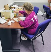 A woman with curly hair wearing a pink long-sleeve shirt working at a table with various items, including papers and cups, in a room with blue chairs.