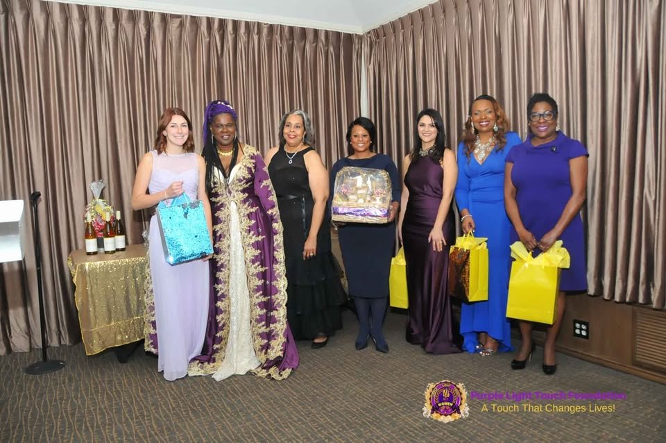 Group of seven women standing indoors, holding gift bags and a gift basket, dressed in formal attire, with a table of wine bottles and candles to the left, and a purple and gold logo with the text "Purple Light Foundation" and "A Touch That Changes L