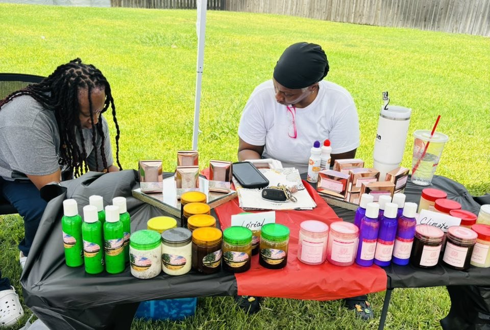 Two women at an outdoor table displaying various skincare and beauty products, including bottles, jars, and boxes, on a grassy area with a fence in the background.