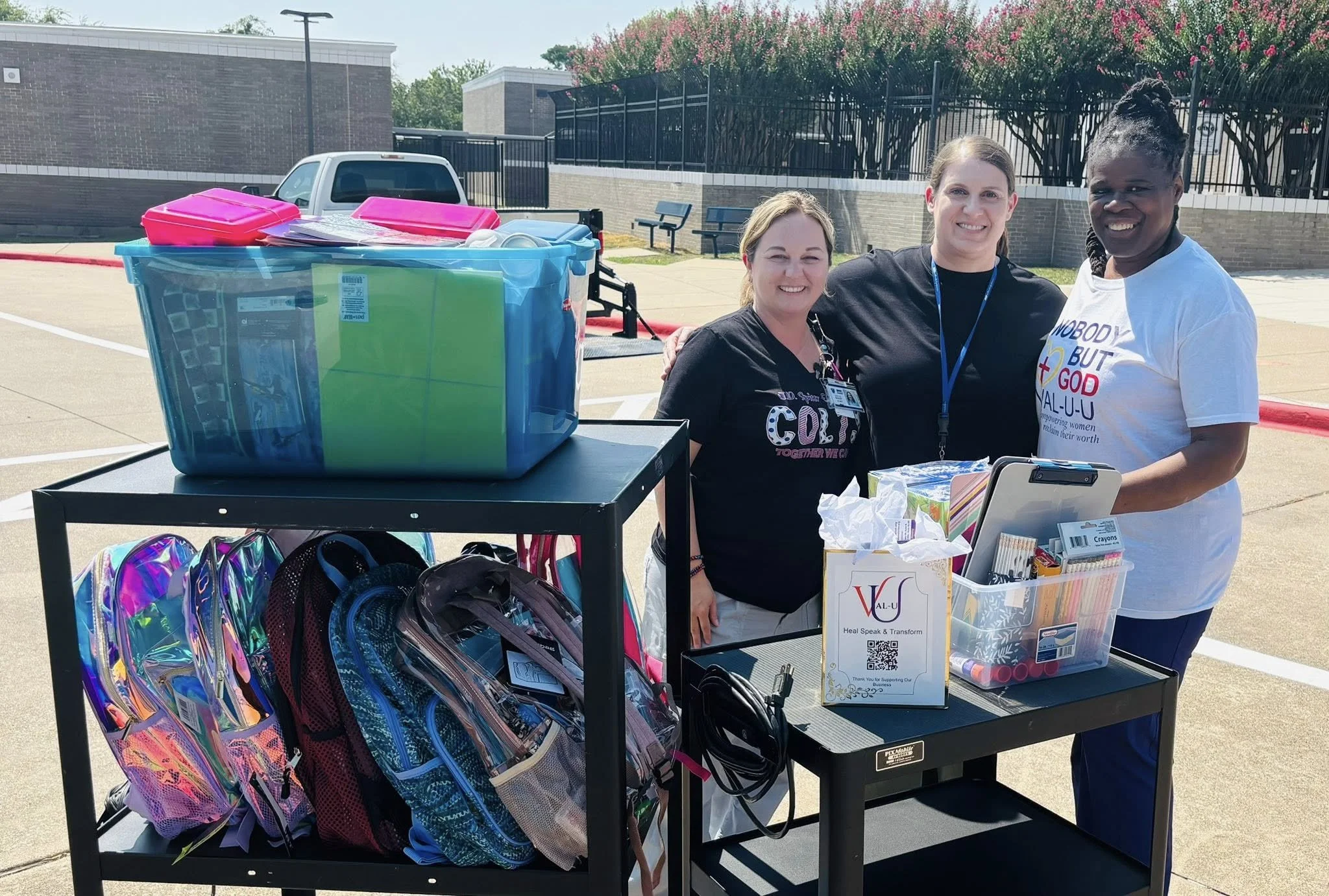 Three women standing behind a table with backpacks, school supplies, and a large plastic container of school items. They are outdoors in a parking lot with a brick building and trees in the background, smiling at the camera.