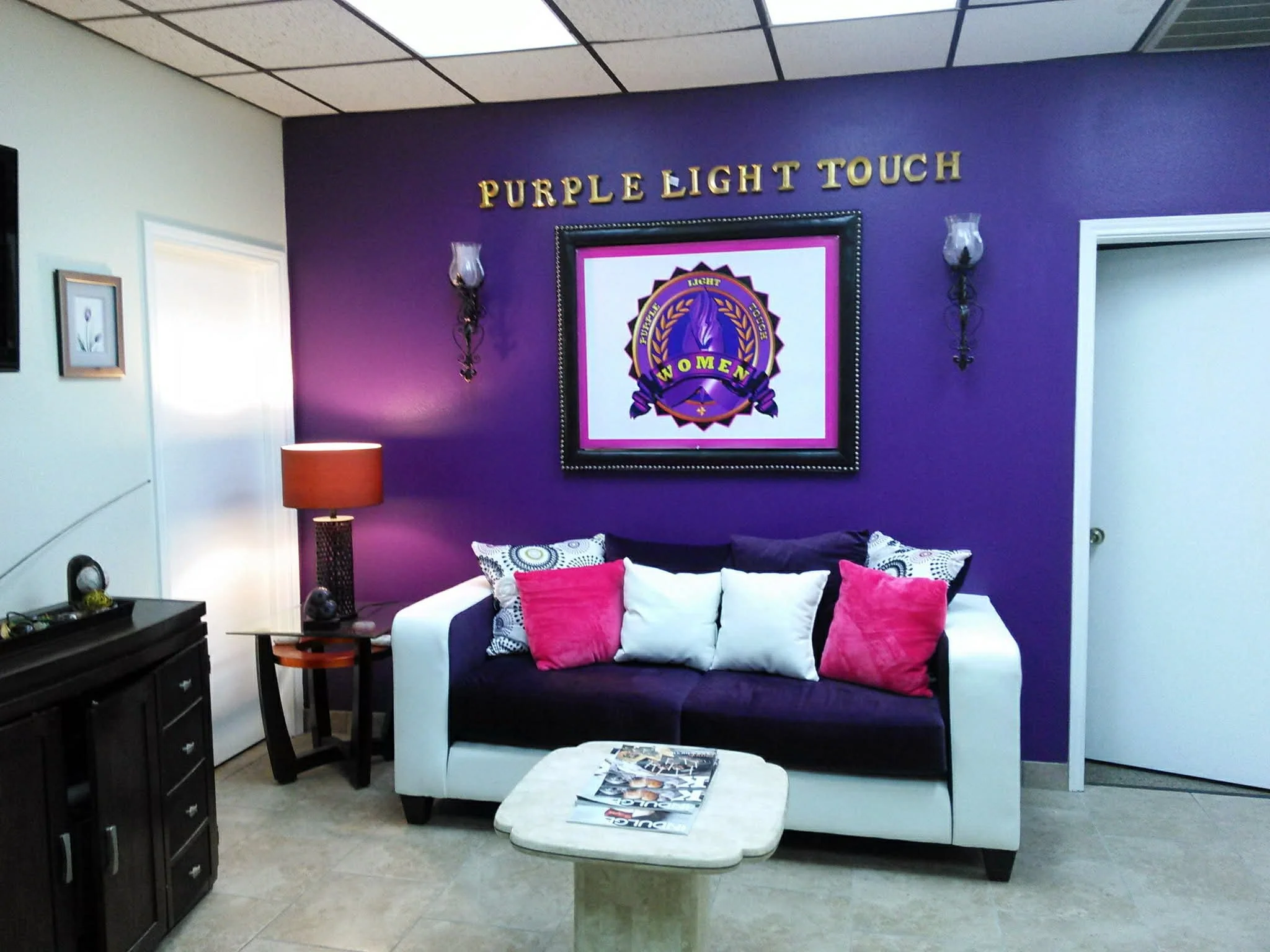 A waiting room with a purple accent wall; the wall has a sign that says 'Purple Light Touch' and a framed emblem with the words 'Women' and a purple flame. There are wall-mounted light fixtures, a white and purple sofa with multiple pink, white, and 