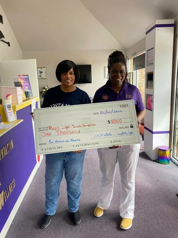 Two women standing indoors holding a giant check for one thousand dollars made out to the Purple Light Touch Foundation.