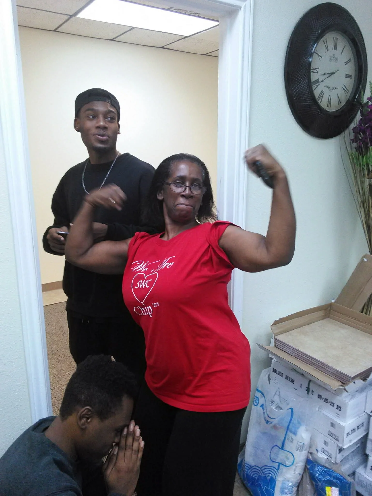 A woman in a red t-shirt flexes her bicep, posing confidently while two young men are nearby. One is standing behind her, and the other is kneeling on the floor with hands clasped in prayer.