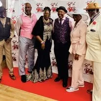 Group of six people standing on a red carpet in front of a white backdrop decorated with pink flowers. They are dressed in formal and semi-formal attire.