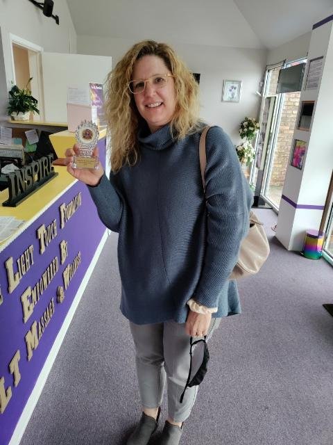 A woman with curly blonde hair and glasses smiling indoors, holding a small award or badge, wearing a blue sweater and gray pants, standing near a reception desk with a purple and yellow sign.