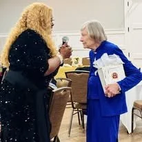 Two women, one young with curly hair in a black dress, and one elderly in a blue suit, smiling and holding a speech or microphone, engaging in conversation in a decorated indoor setting.