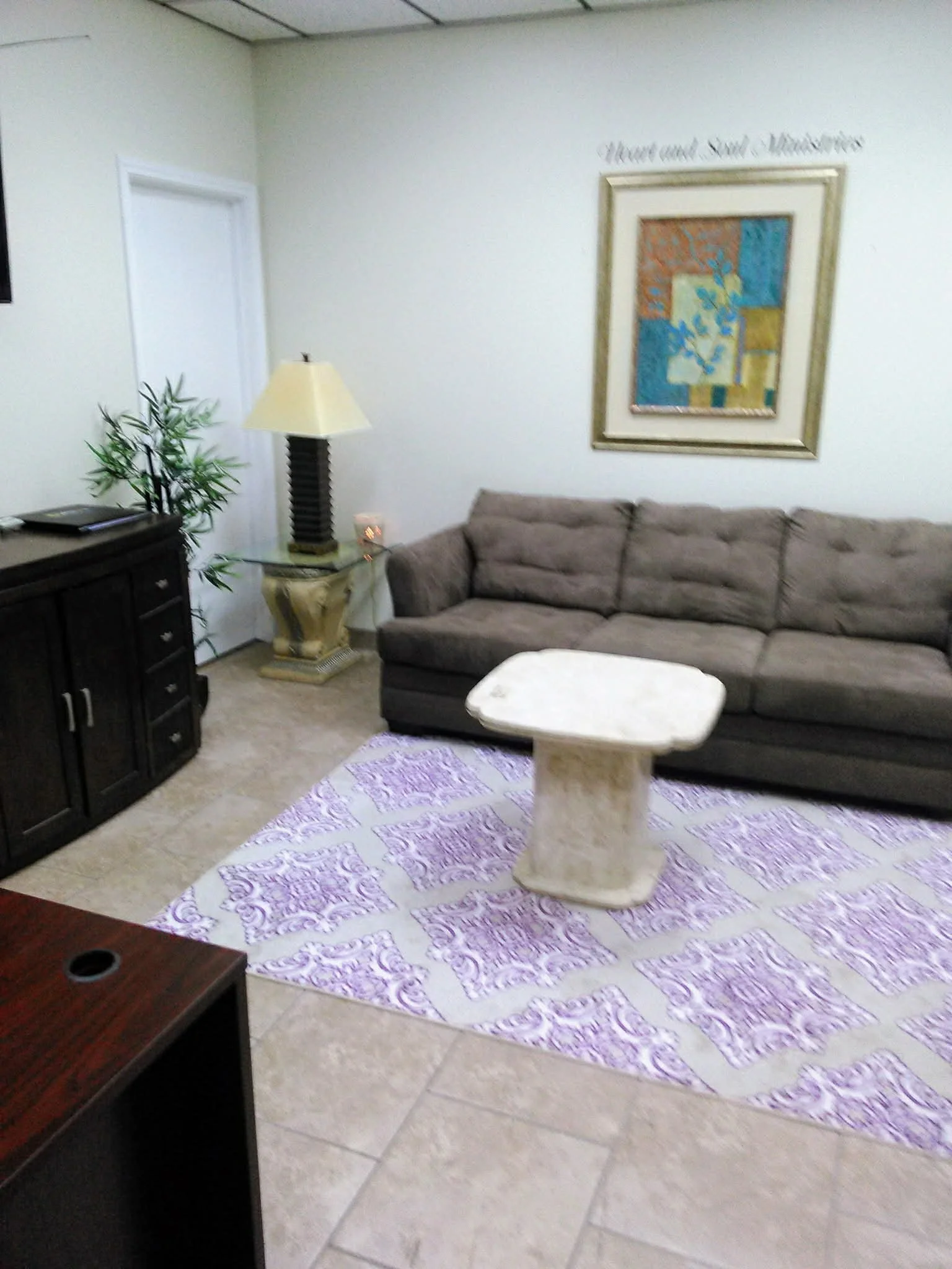 Living room with a brown sofa, a purple and white patterned rug, a white marble coffee table, a black cabinet, a tall lamp, and a framed artwork on the wall.