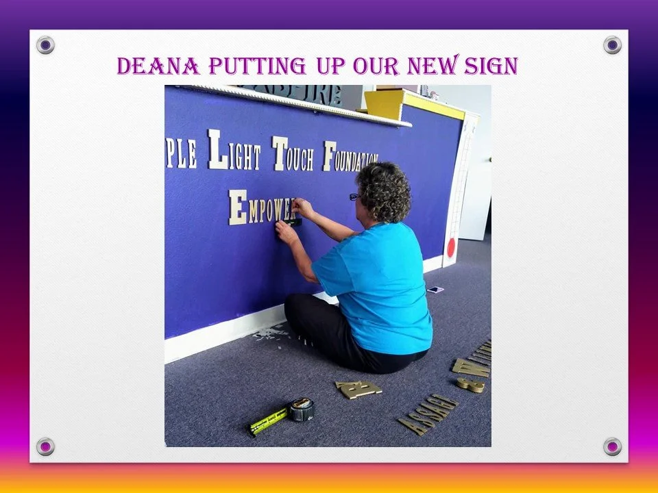 A woman in a blue shirt sitting on the floor attaching letter cutouts to a blue wall, with a new sign that reads 'Deanna Putting Up Our New Sign.'