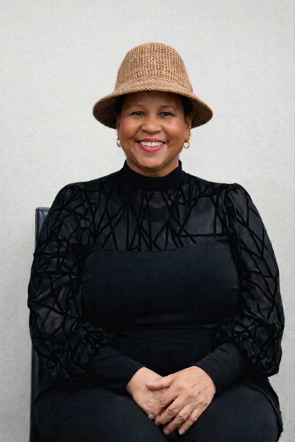 A smiling woman with short hair, wearing a tan woven hat, gold hoop earrings, a black patterned top with sheer sleeves, and sitting against a plain white wall.