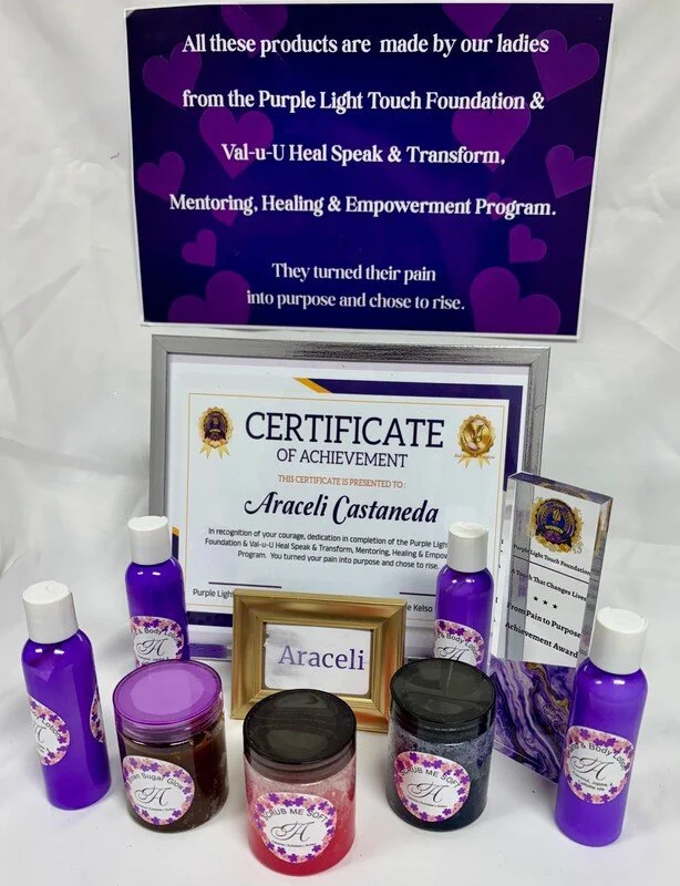 A display of purple bottles, jars, and a framed certificate of achievement with a purple, heart-themed sign above that mentions products made by women from the Purple Light Touch Foundation and Val-u-U Heal Speak & Transform, emphasizing empowerment 