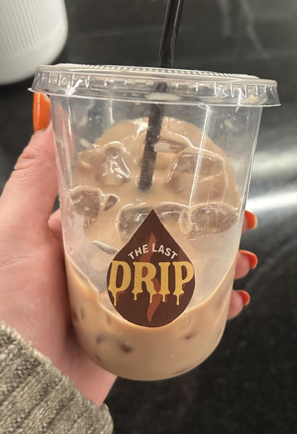 A clear plastic cup filled with iced coffee with milk, ice cubes, a black straw, and a sticker that says "The Last Drip."