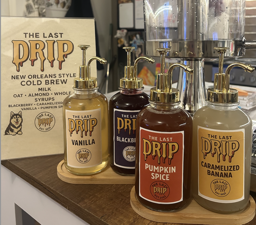 Four bottles of flavored syrup, labeled Vanilla, Blackberry, Pumpkin Spice, and Caramelized Banana, are placed on a wooden board. A menu or sign for The Last Drip New Orleans style cold brew is visible in the background.