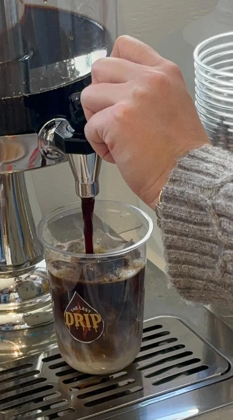 A person in a gray sweater is pouring black coffee from a coffee machine into a clear plastic cup labeled "The Last Drip".