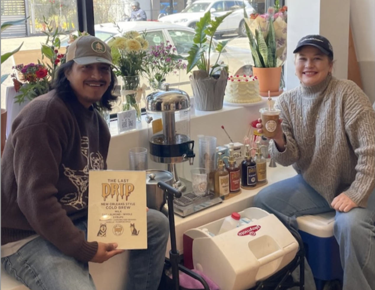 Two people smiling and sitting at a table inside a cafe, surrounded by flowers, with a variety of drinks, syrups, and a menu. The person on the left is holding a menu, and the person on the right is holding a drink with a straw. They are dressed casually, and the scene looks cheerful and cozy.