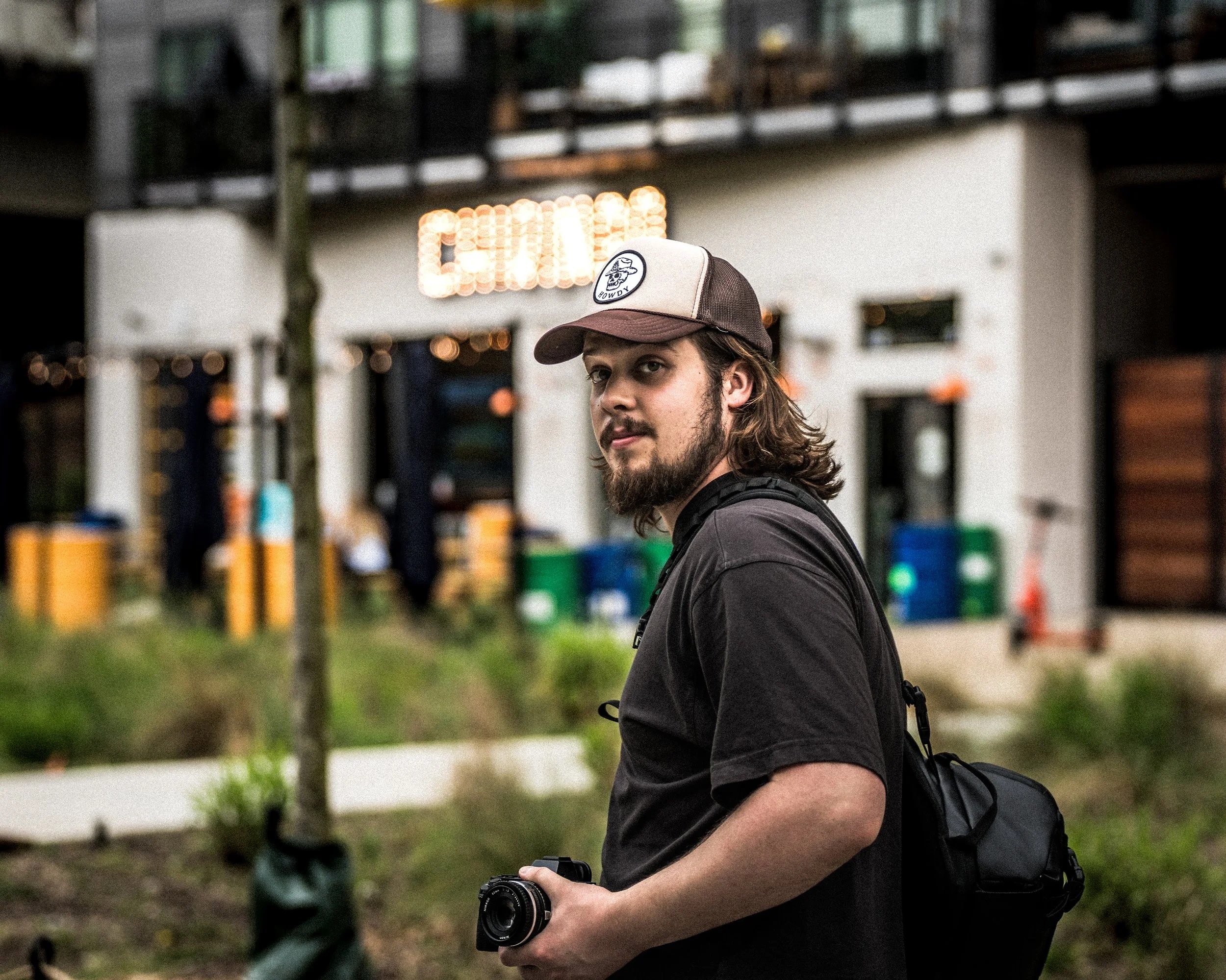 A man with long hair and a beard wearing a baseball cap, black T-shirt, and backpack holding a camera, standing outdoors in front of a building with a blurred background.