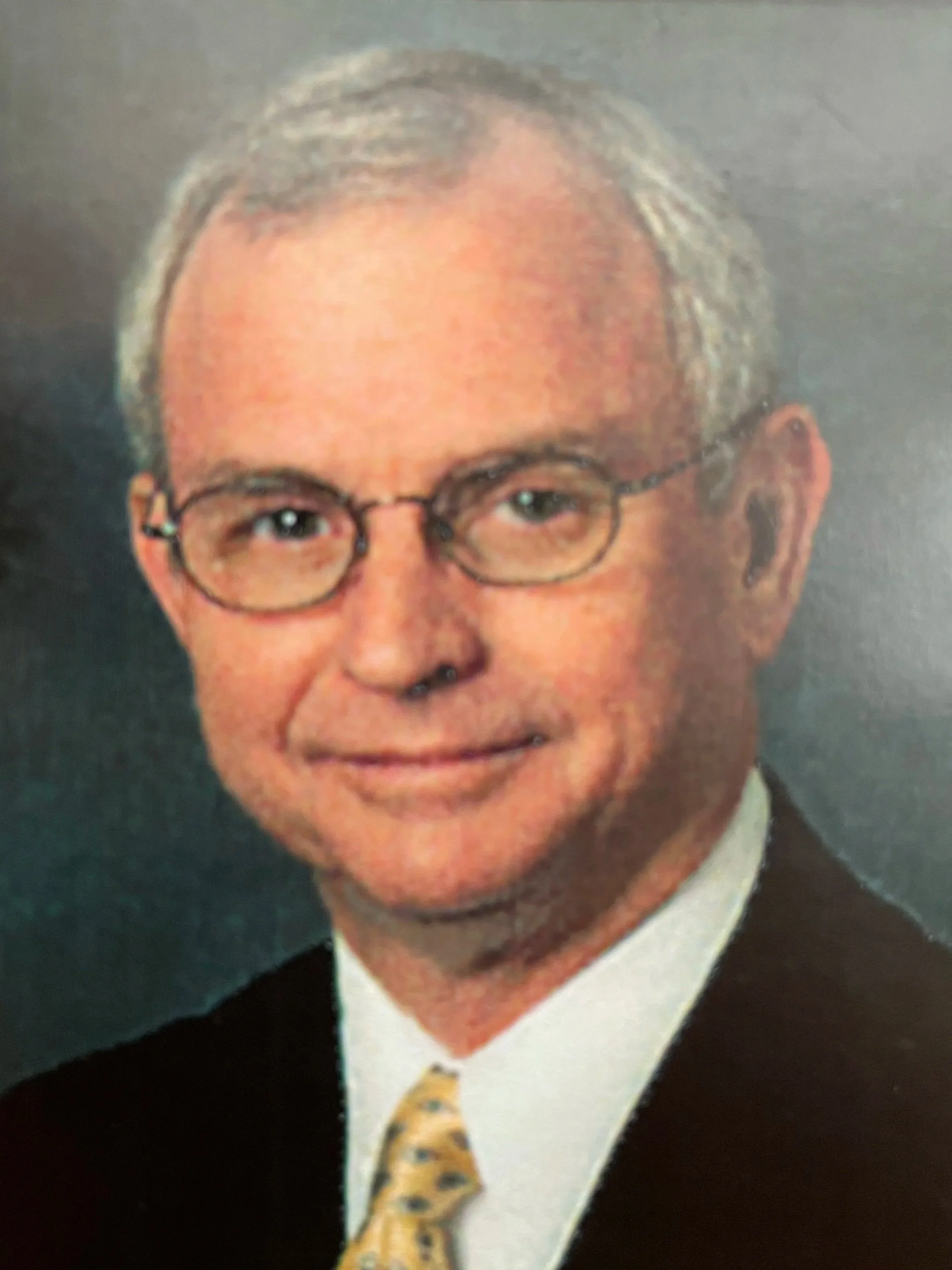 A headshot of an older man with gray hair, glasses, wearing a suit and a patterned tie.