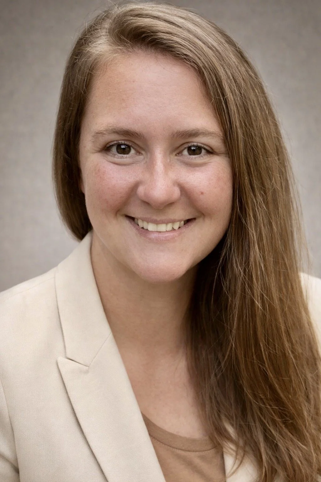 A woman with long, reddish-brown hair, smiling, wearing a beige blazer and a light brown top, posed against a neutral background.