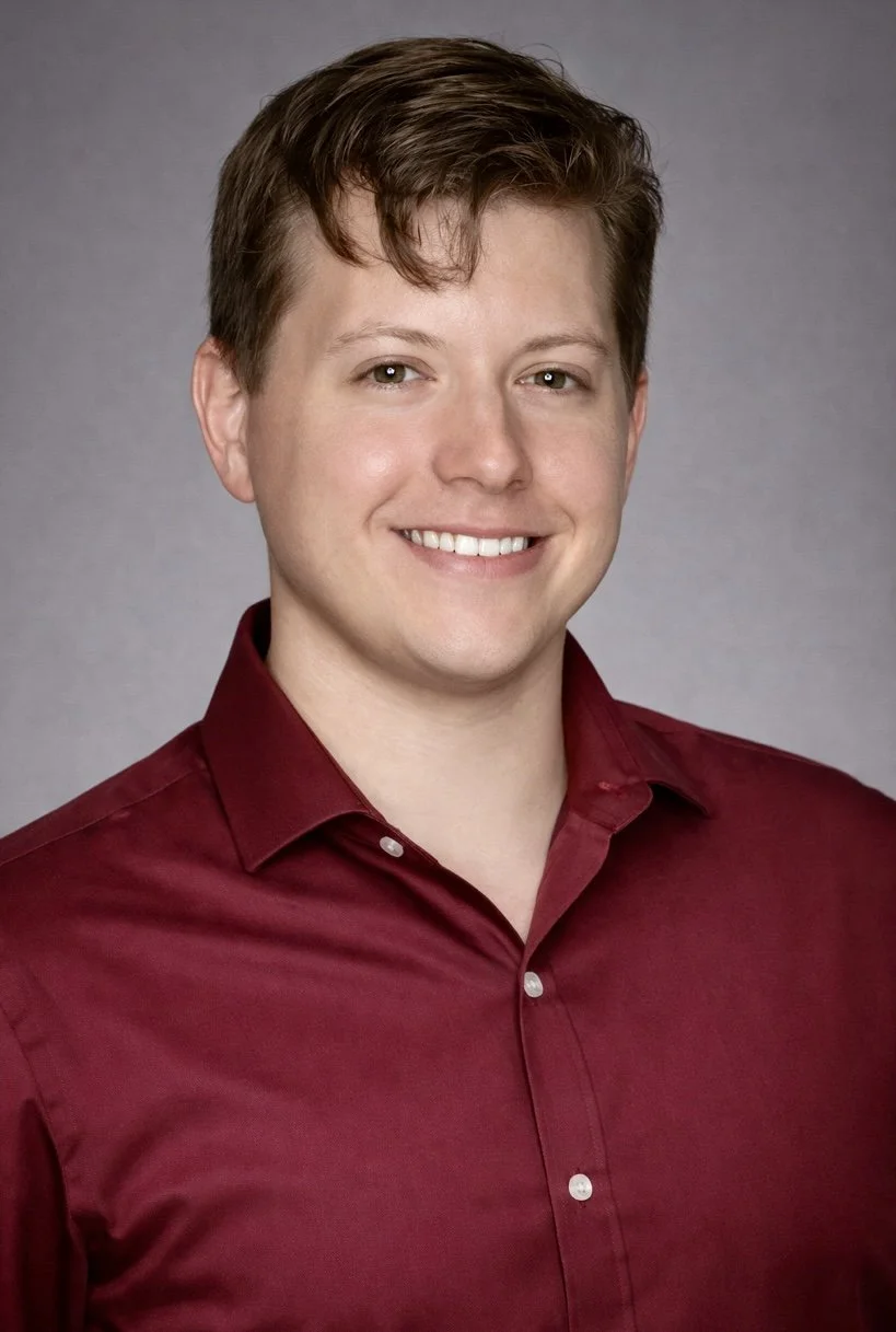 A young man with short brown hair and light skin, wearing a red button-up shirt, smiling against a plain gray background.