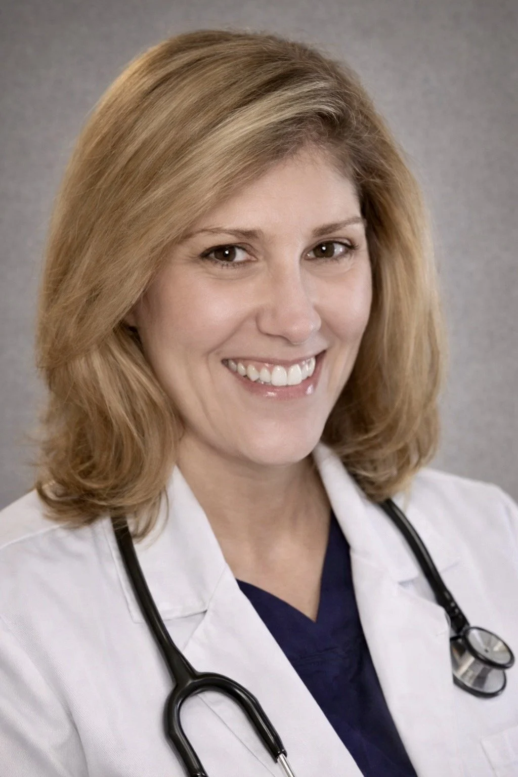 A female doctor with shoulder-length blonde hair smiling, wearing a white coat and a stethoscope around her neck.
